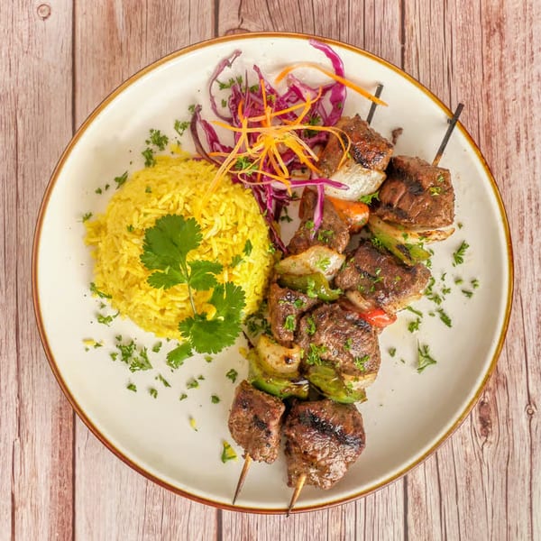 Two beef and vegetable skewers with yellow rice, garnished with cilantro and shredded cabbage.