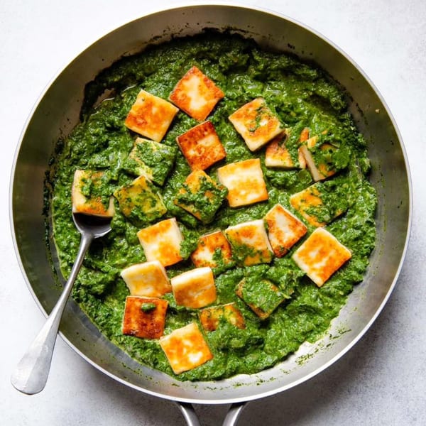 A skillet of vibrant green spinach curry with golden-brown paneer cheese cubes and a spoon.