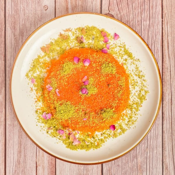 A vibrant orange Mediterranean dessert, likely kunafa, topped with green pistachios and pink rose petals.
