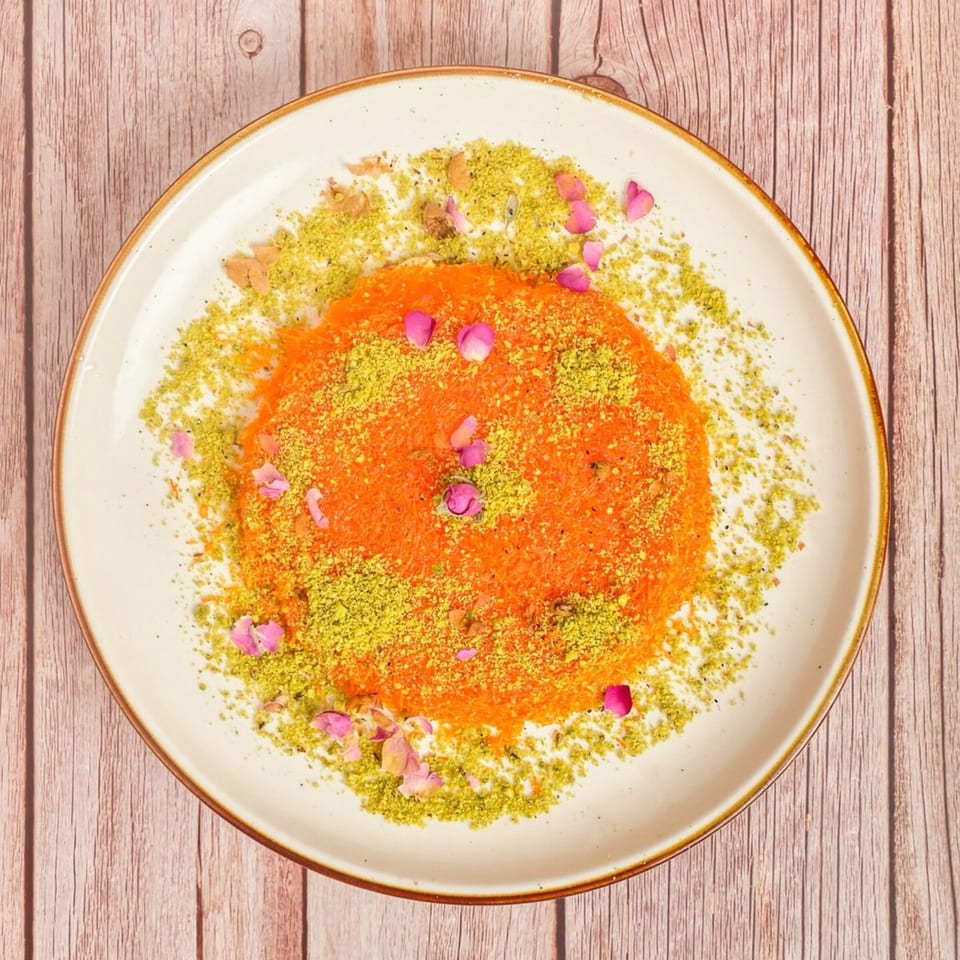 A vibrant orange Mediterranean dessert, likely kunafa, topped with green pistachios and pink rose petals.